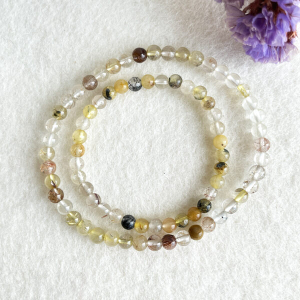 Two strands of multicolored beaded bracelets laid out in circles on a white textured background, with a blurred purple flower in the top right corner.