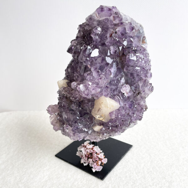 A large amethyst geode on a black stand with a cluster of small, pale pink flowers at its base, set against a white background.