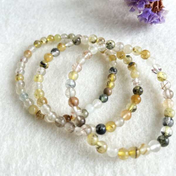 Three beaded bracelets with various hues of rutilated quartz beads, arranged on a white fuzzy surface with a purple dried flower in the background.