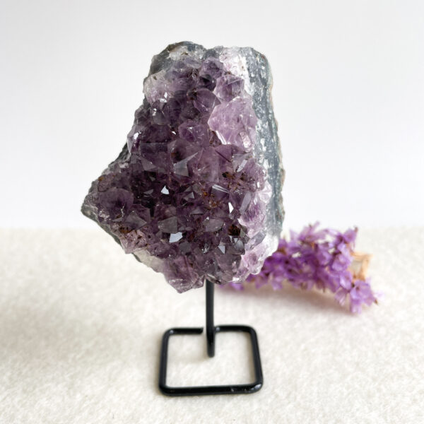 A large amethyst geode on a black metal stand with small purple flowers lying next to it on a white surface.