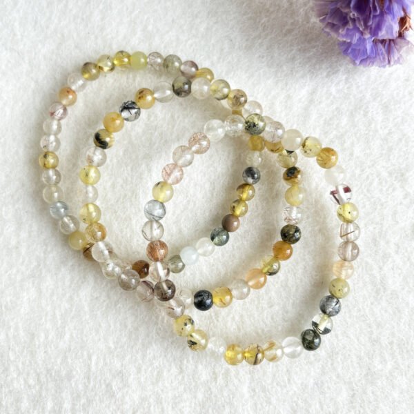 Three strands of multicolor beaded bracelets laid out in overlapping loops on a white textured surface with a blurred purple flower in the corner.
