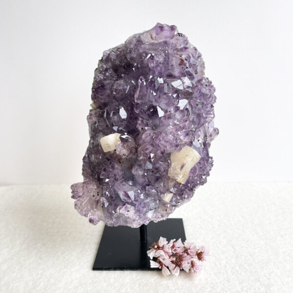 A large amethyst geode on a black stand with some pink dried flowers at its base, set against a white background.
