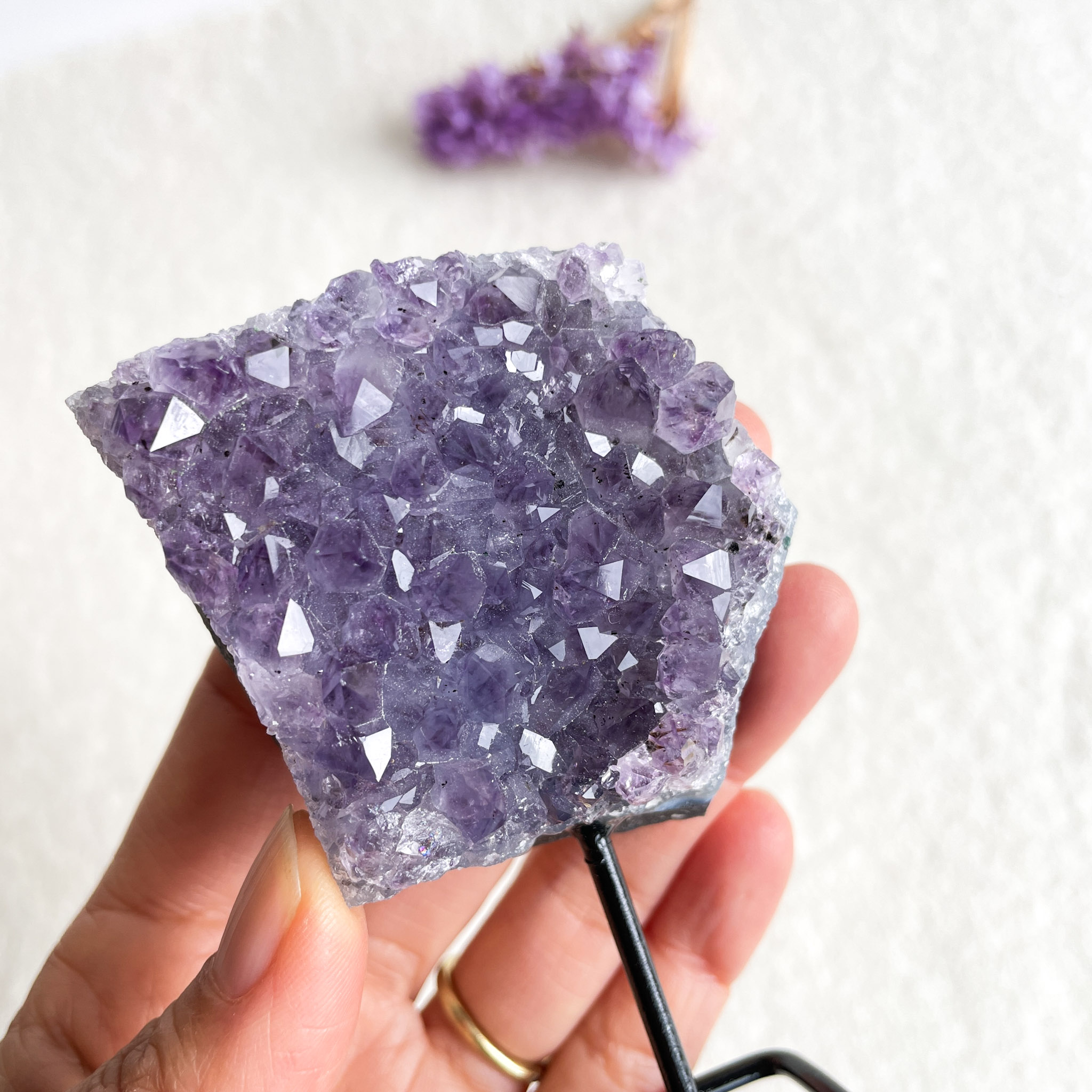 A person's hand holding a piece of purple amethyst crystal against a light background, with a blurred small purple flower in the distance.
