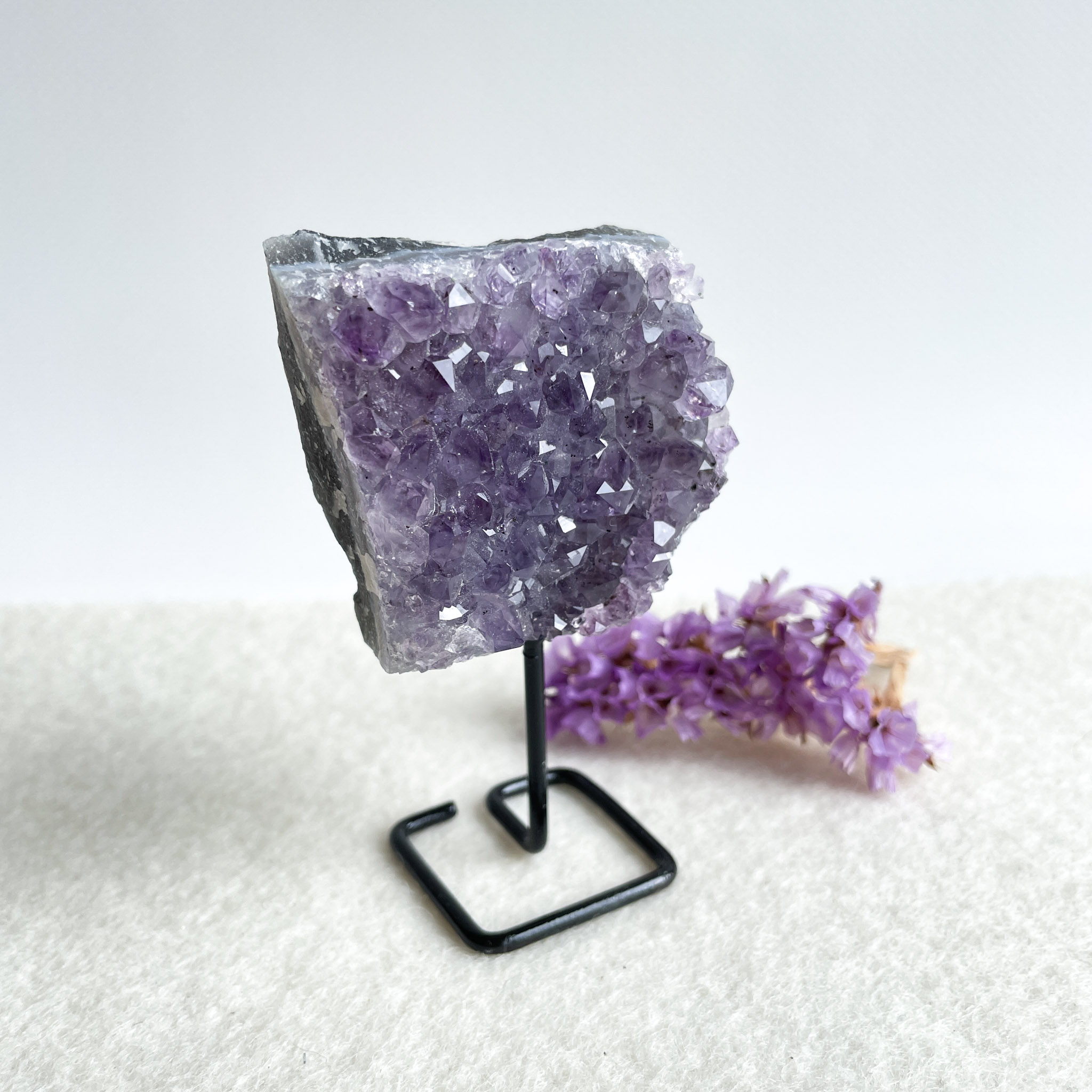 A vibrant purple amethyst geode displayed on a black metal stand, with a small bundle of purple dried flowers lying next to it against a white background.