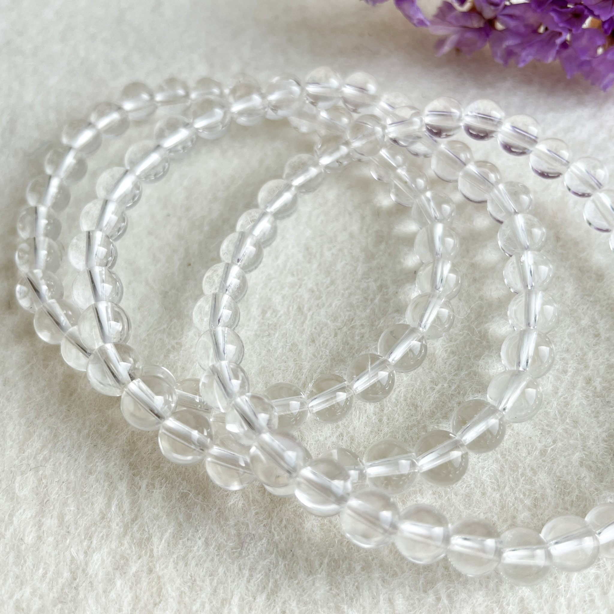 Three strands of clear glass beads on a white surface with a hint of purple flowers in the top right corner.