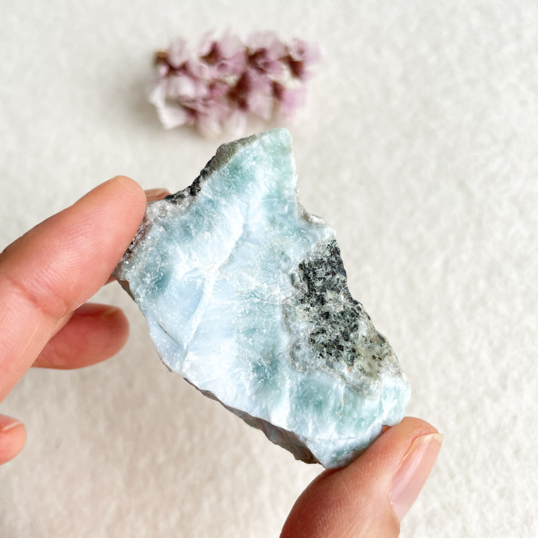 A hand holding a piece of light blue translucent raw mineral with natural edges against a white background, with small pink flowers blurred in the distance.