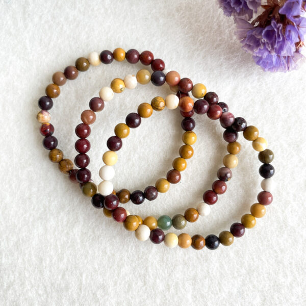 Three intertwined beaded bracelets made of multicolored stones laid out on a white textured surface with a blurred purple flower in the corner.