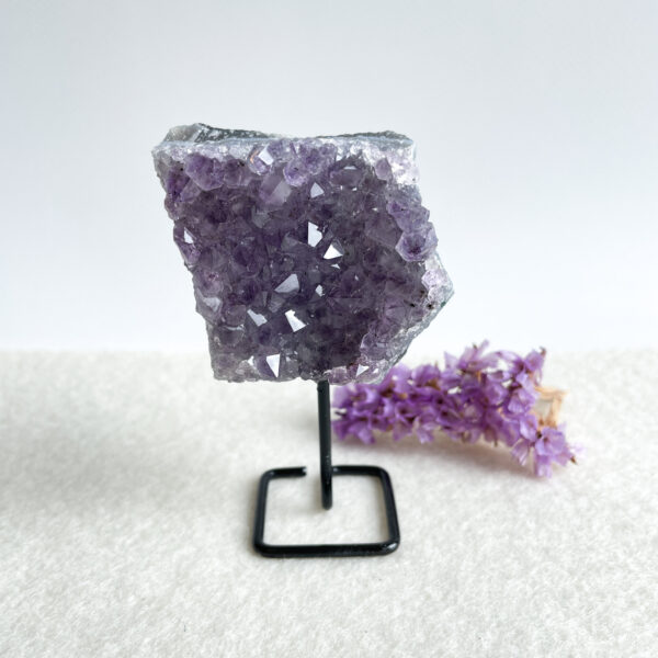 A large amethyst crystal on a black metal stand with a cluster of small purple flowers lying next to it on a white textured surface.