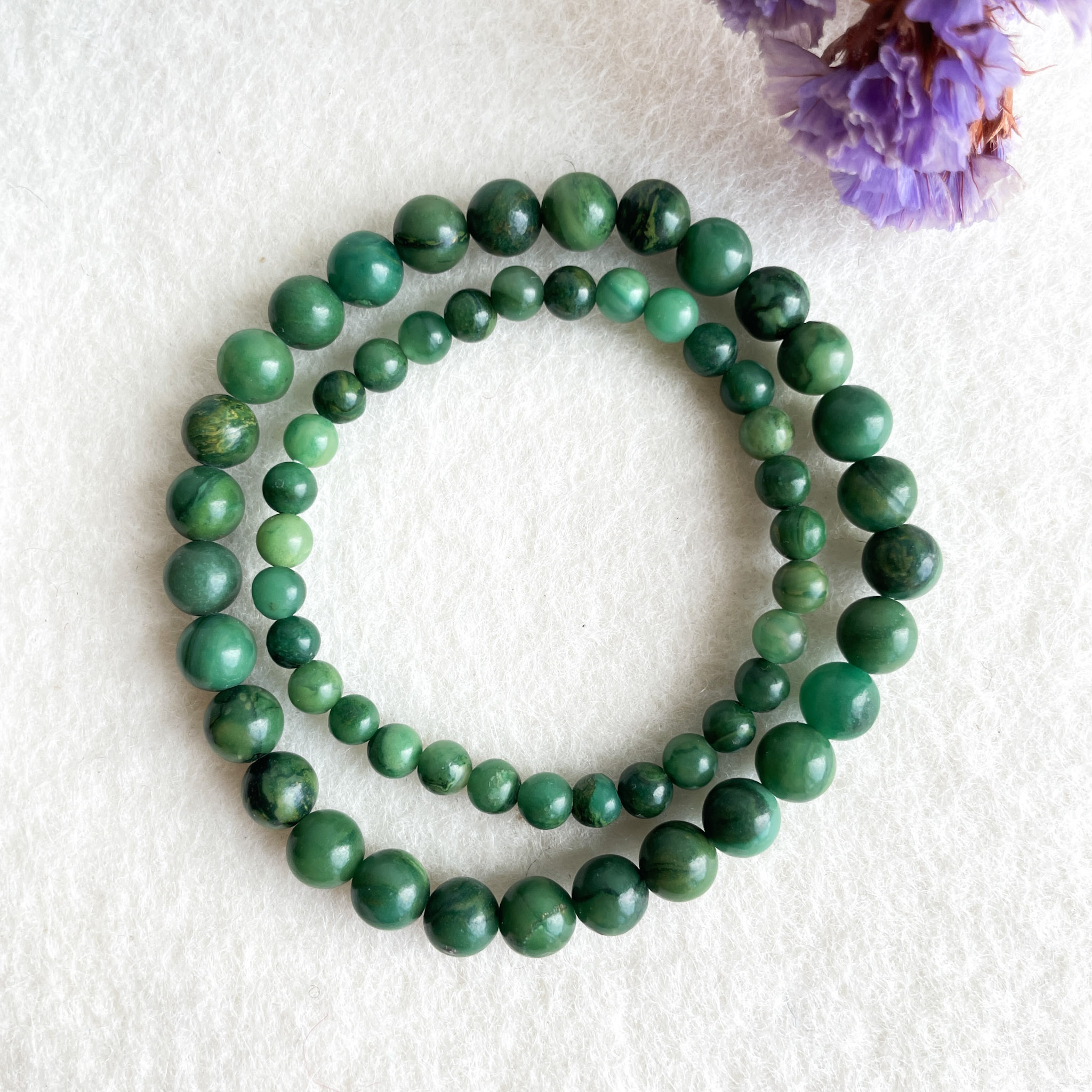 A green stone bead necklace arranged in a spiral shape on a white background with a blurred purple flower in the corner.