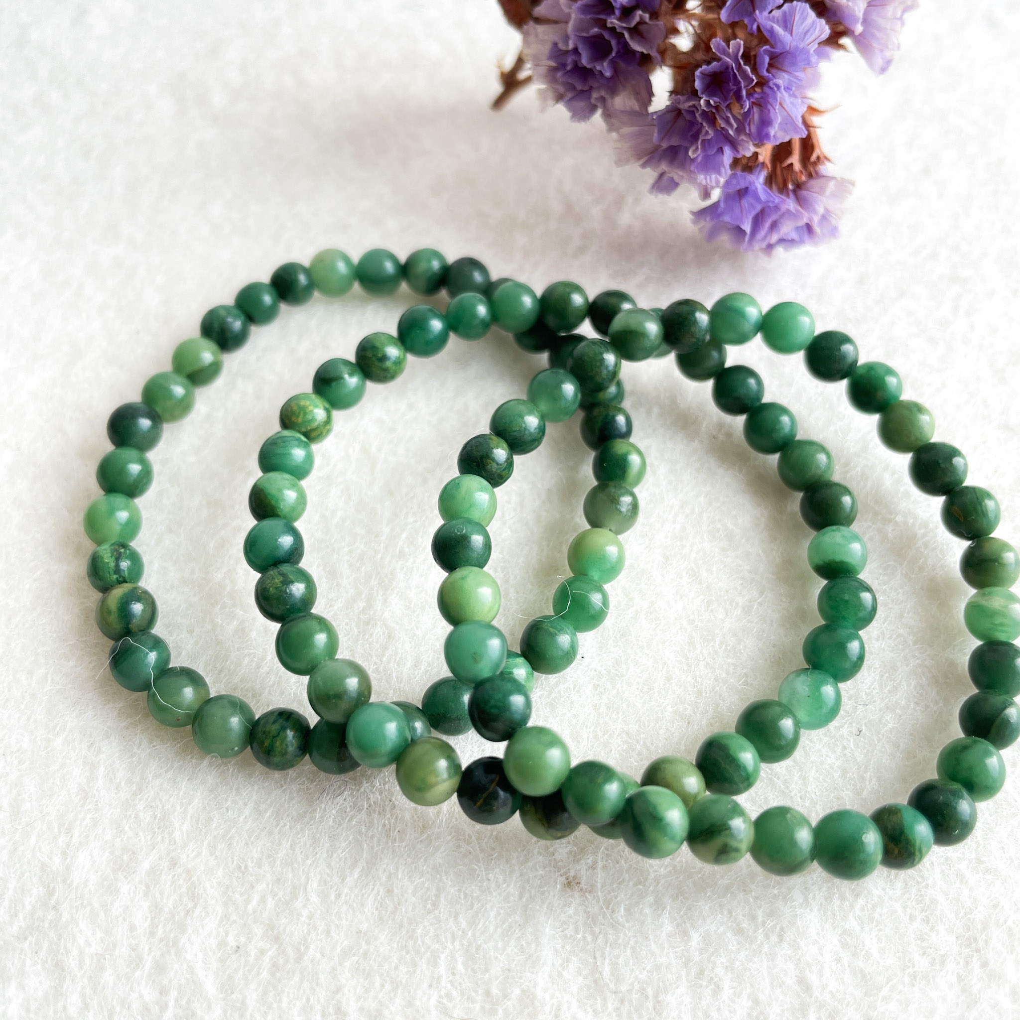 Three strands of green jade beads laid on a white surface with blurred purple flowers in the background.