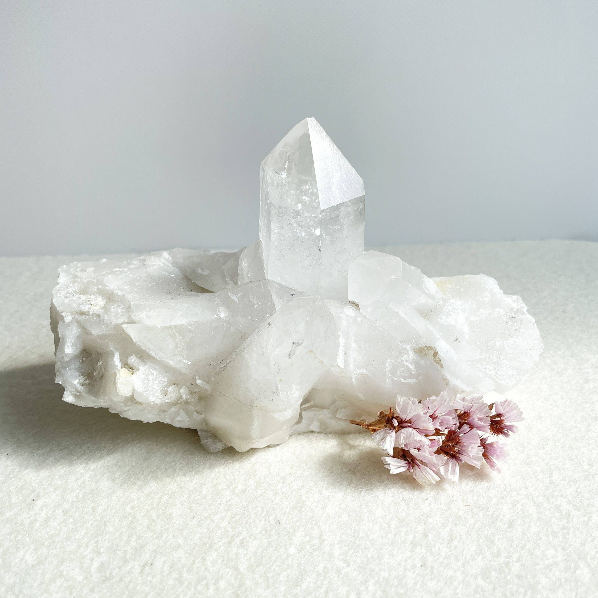 A cluster of clear quartz crystals with a prominent central point, accompanied by small pink blossoms on a white surface with a pale background.