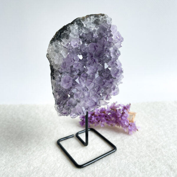 An amethyst geode on a black metal stand with a small bunch of purple flowers lying next to it on a white surface.
