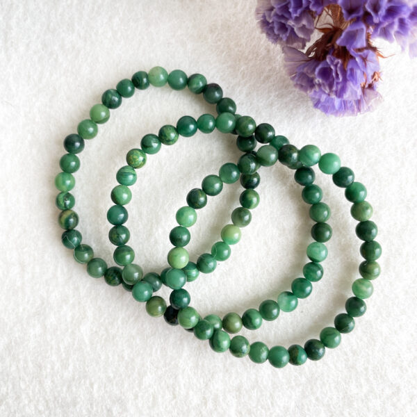 Three strands of green jade bead bracelets are laid out on a white surface, with a blurred purple dried flower in the upper right corner.