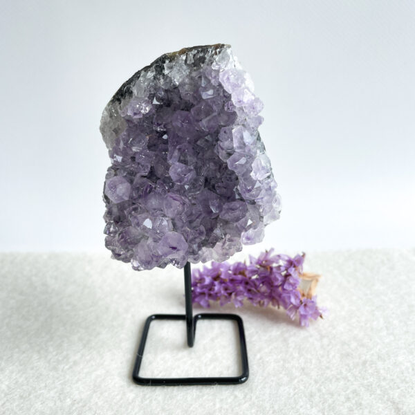 A large amethyst geode with deep purple crystals on a black metal stand, with a sprig of lilac flowers to its right, on a white textured surface against a light background.