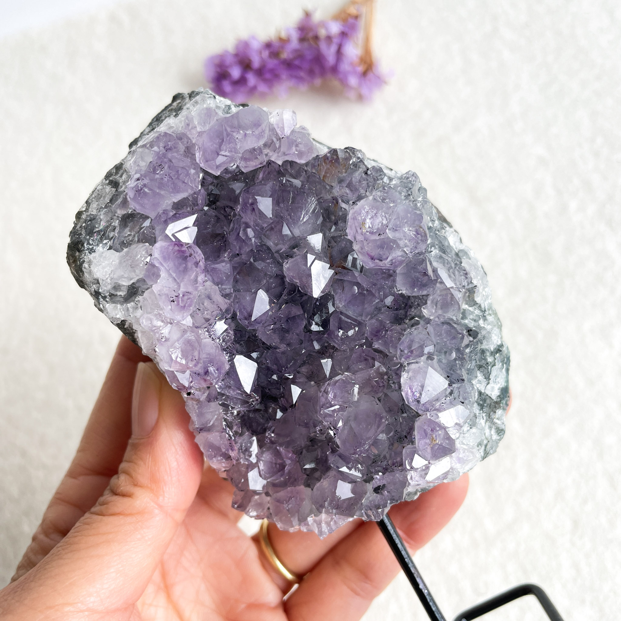 A person holds a large amethyst geode with well-defined purple crystals against a light background, with a small purple flower in the top right corner.