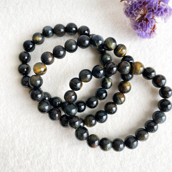A string of shiny, black tiger's eye stone beads arranged in spiral loops on a white textured surface with a small bundle of dried lavender flowers in the top right corner.