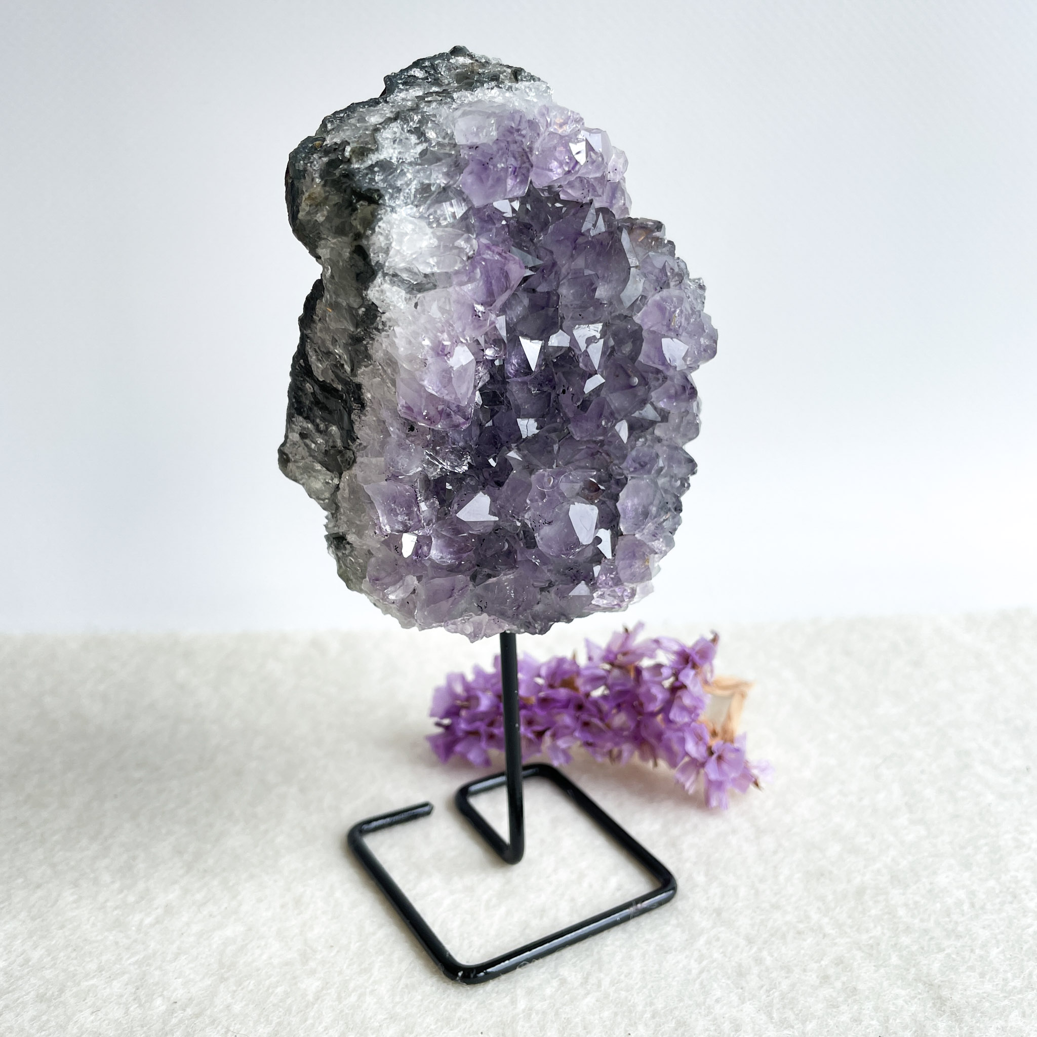 A large amethyst geode on a black metal stand with small purple flowers beside it, placed on a textured white surface against a white background.