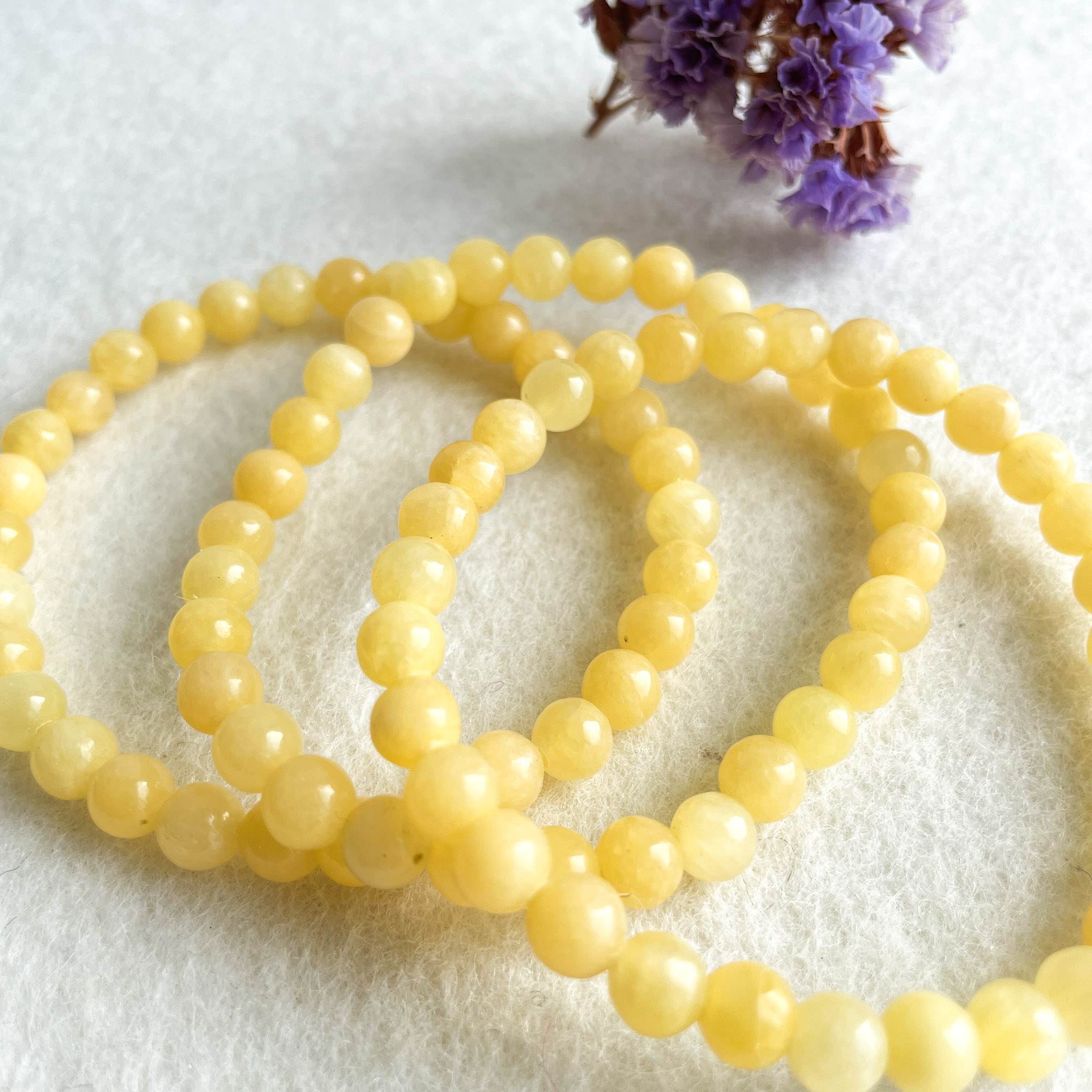 Three strands of yellow beaded bracelets laid out on a white textured surface with a small bunch of purple dried flowers at the top right corner.