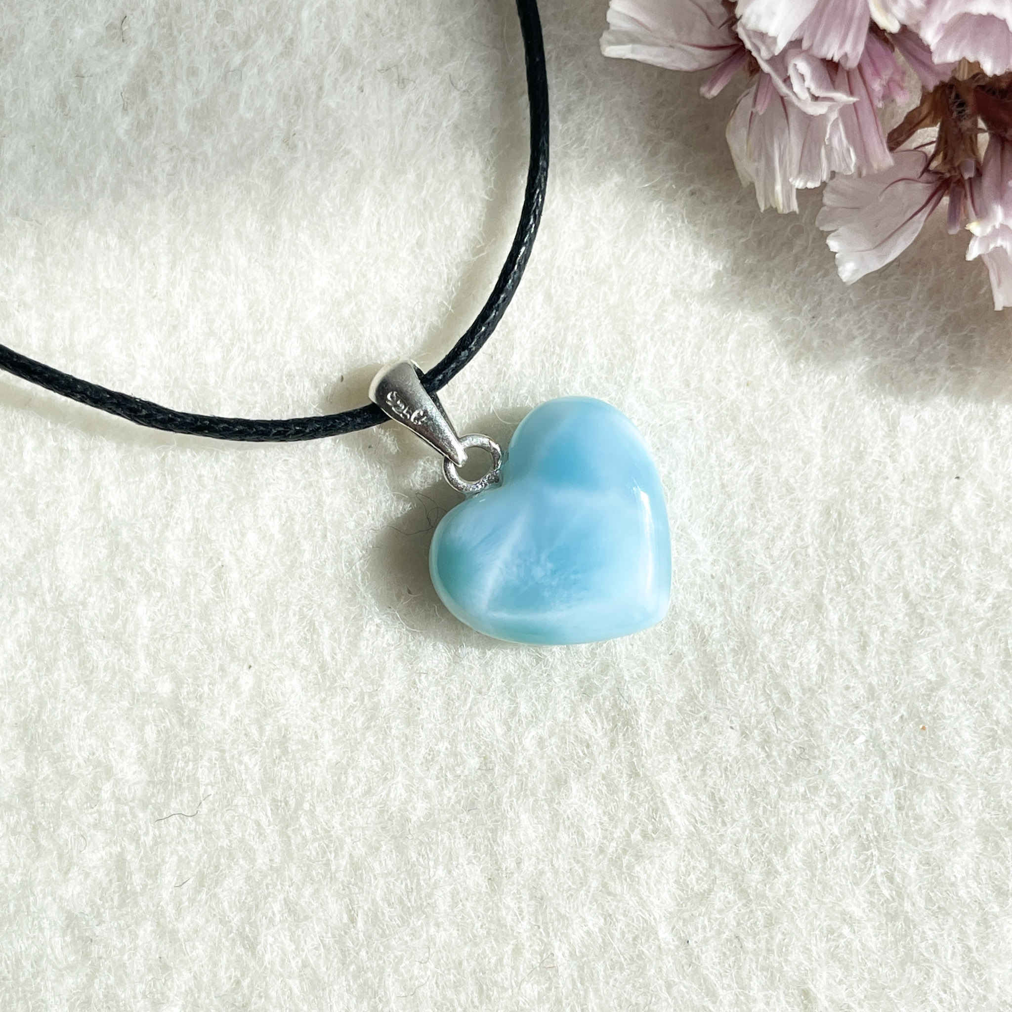 A light blue heart-shaped pendant on a black cord necklace, lying on a white textured background next to pink flowers.