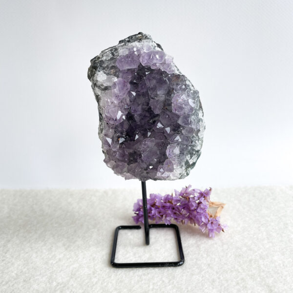 A large amethyst geode on a metal stand with small purple flowers lying next to it on a white surface against a white background.