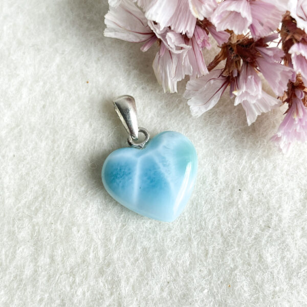 A blue heart-shaped pendant with a silver bail on a white textured surface, accompanied by pink flower petals.