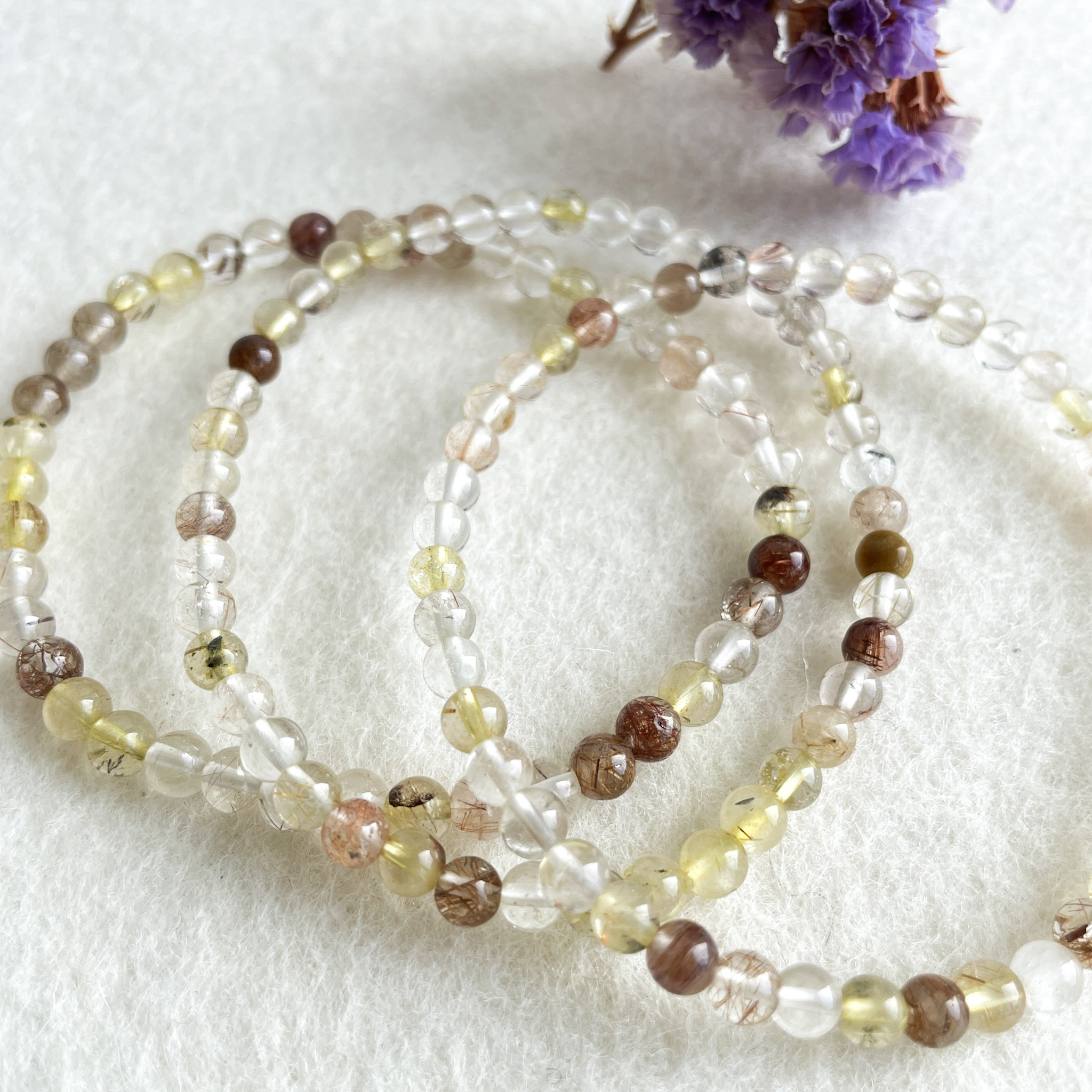 Three strands of crystal bead necklaces with varying shades of brown, yellow, and clear beads, displayed on a white textured background with purple dried flowers partially visible in the background.