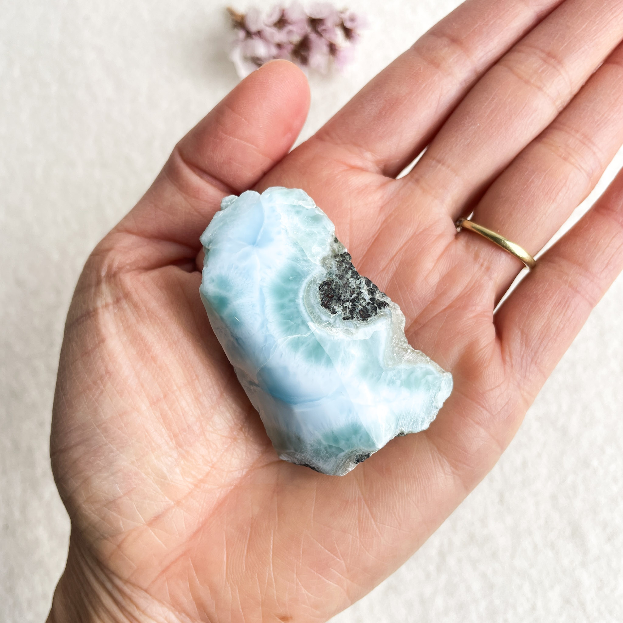 A person's open hand displaying a translucent blue larimar stone with patches of white and gray, with a blurred background of small purple flowers and a light surface.