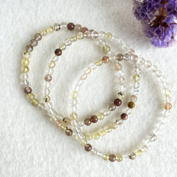 A set of beaded crystal bracelets in various shades of brown, yellow, and clear laid out on a white fuzzy surface, with a portion of a purple flower visible in the top right corner.