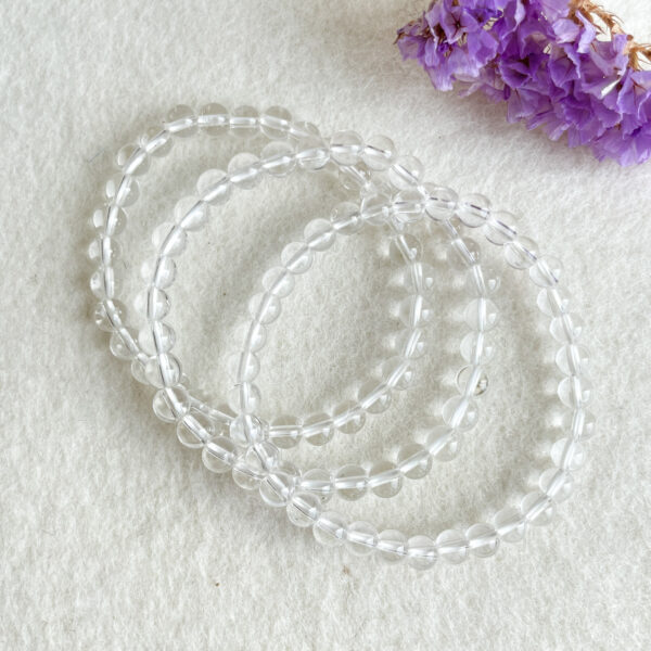 Three strands of clear crystal bead bracelets on a white textured background with a bundle of purple dried flowers in the top right corner.