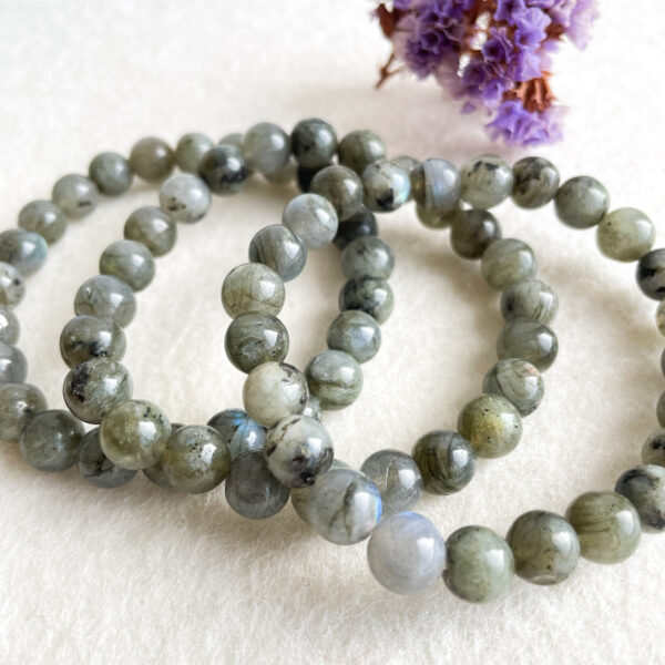 Three strands of polished labradorite bead bracelets with varying shades of grey and blue, displayed on a neutral background with a small dried flower in the corner.
