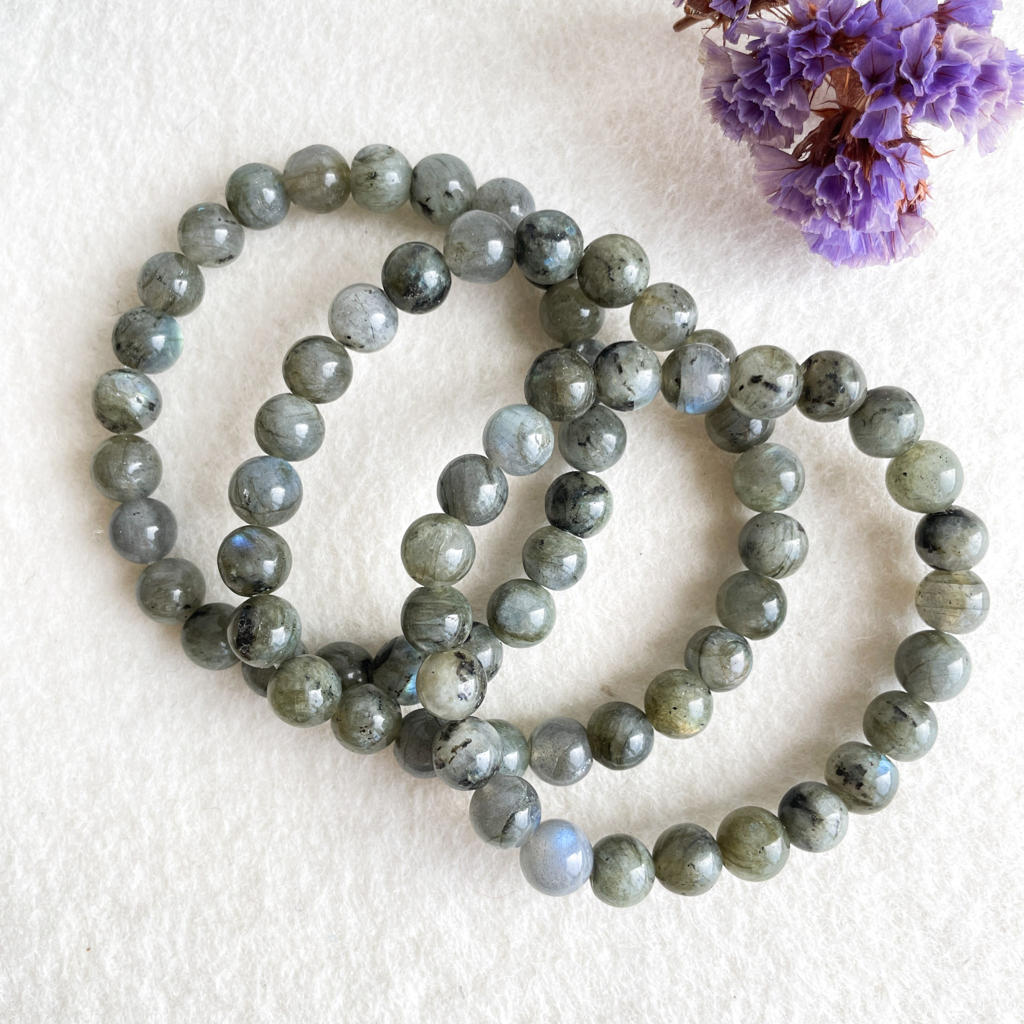 Two strands of labradorite beaded bracelets on a white textured background with a purple dried flower in the top right corner.