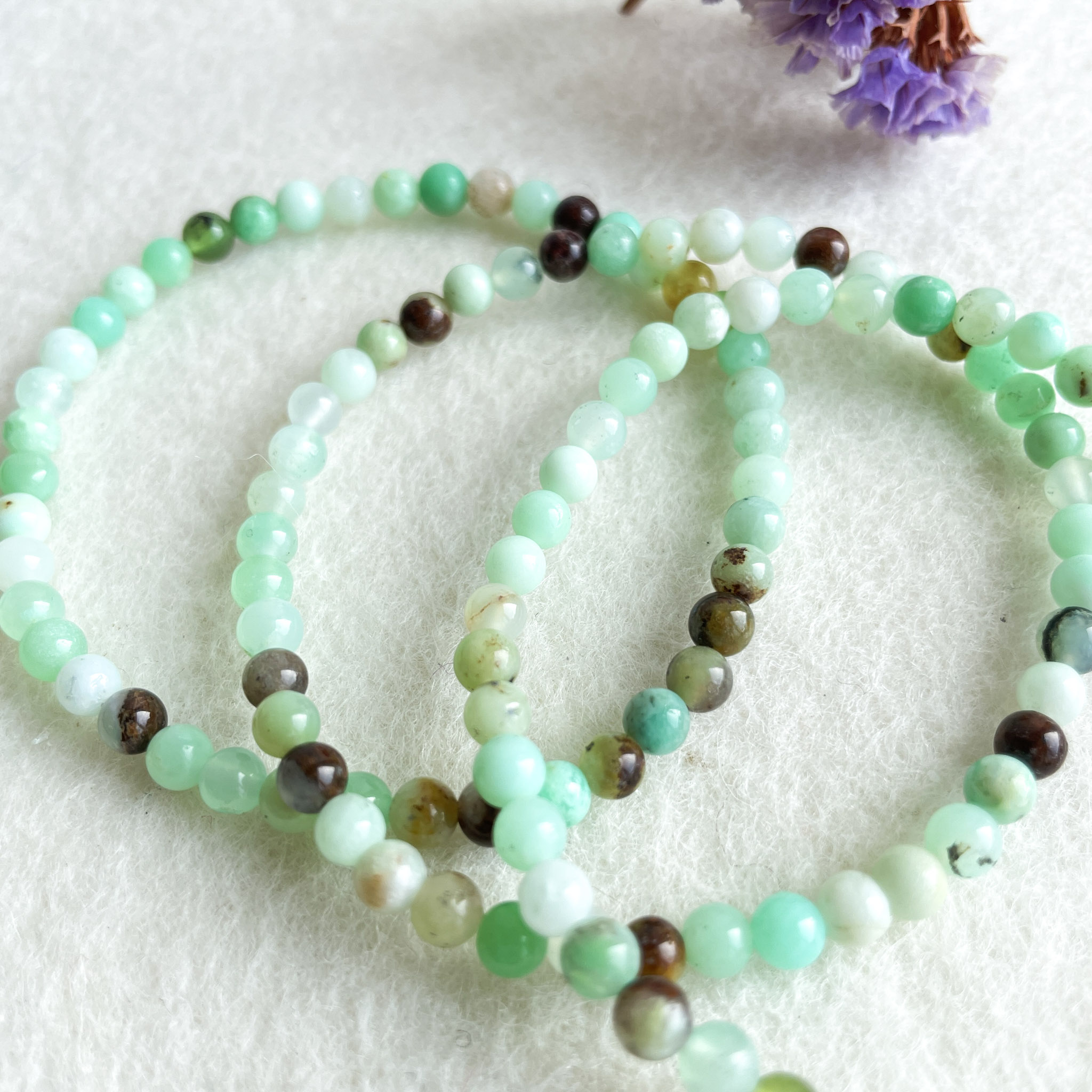 A set of two beaded necklaces with varying shades of green beads, some of which are translucent, interspersed with occasional brown beads, laid out on a white fibrous background next to a small purple flower.