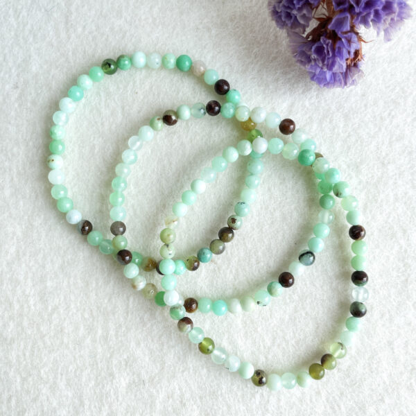 A jade bead necklace in various shades of green laid out on a textured white surface next to purple dried flowers.