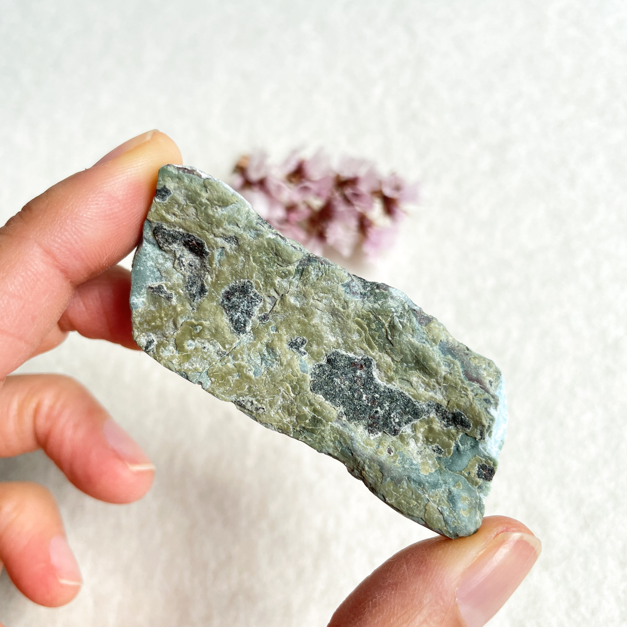 A person holding a slab of green and black rock with small, pink crystal formations in the blurry background.