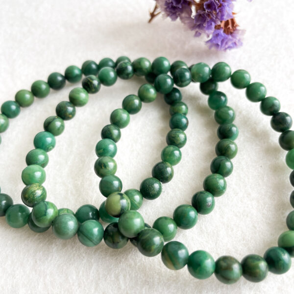 A necklace of polished green stone beads laid out in a loop on a white surface, with blurred purple flowers in the background.