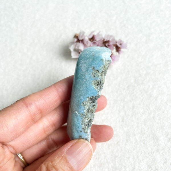 A close-up photo of a person's fingers holding a polished larimar stone with a textured gray inclusion, with small purple flowers in the background on a white surface.