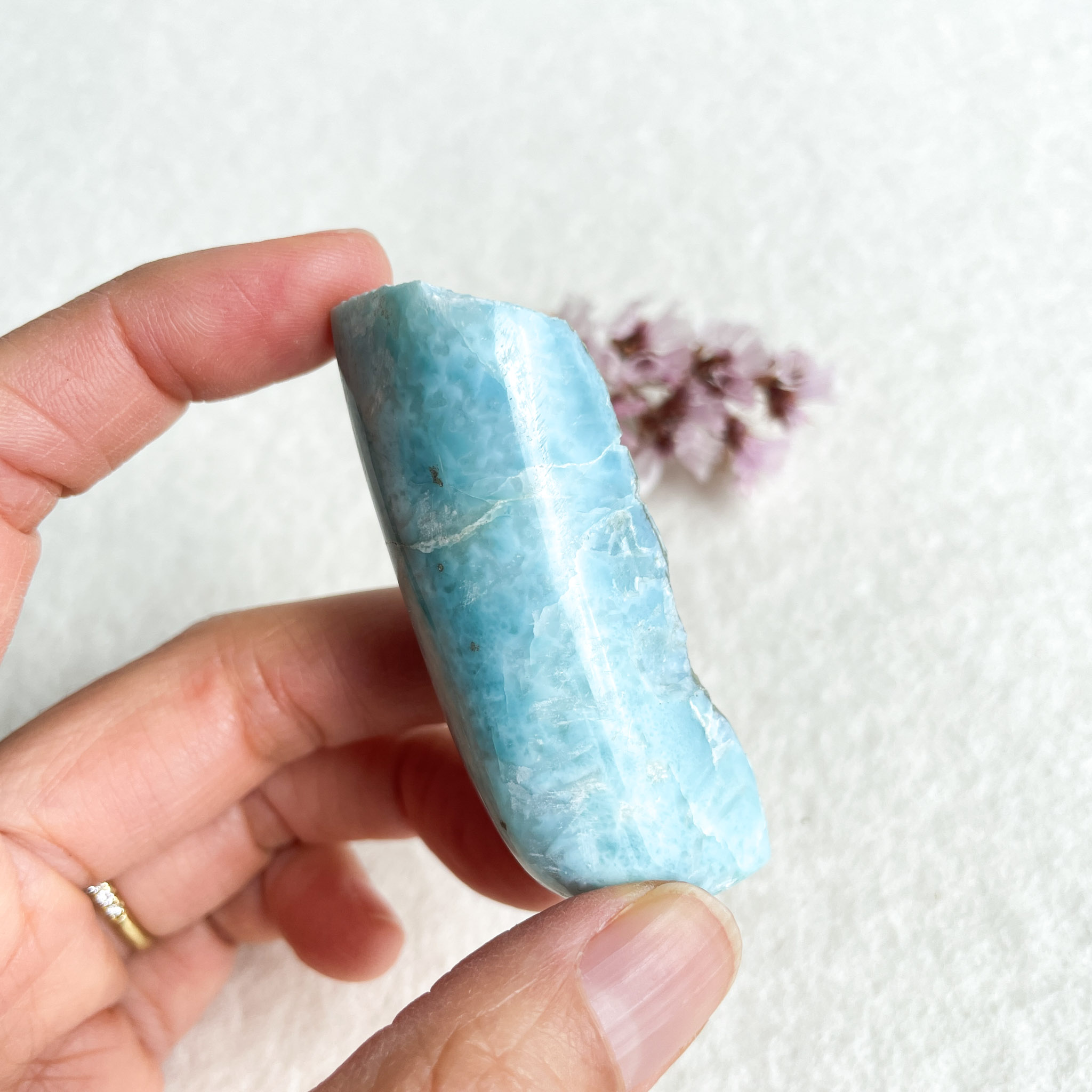A person's fingers holding a polished light blue aquamarine crystal with natural fractures, against a white background with a blurred cluster of small purple flowers in the corner.