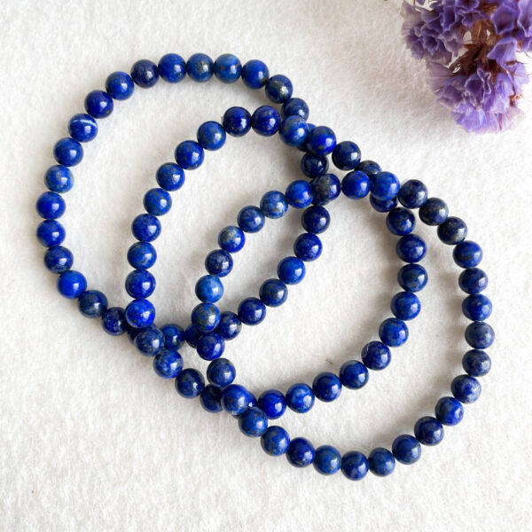 Three strands of lapis lazuli bead necklaces arranged in spirals on a white textured background with a dried purple flower in the corner.