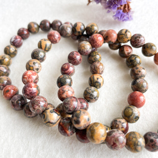 A close-up of a beaded necklace with multicolored marbled beads on a white fuzzy surface, with a small purple flower blurred in the background.