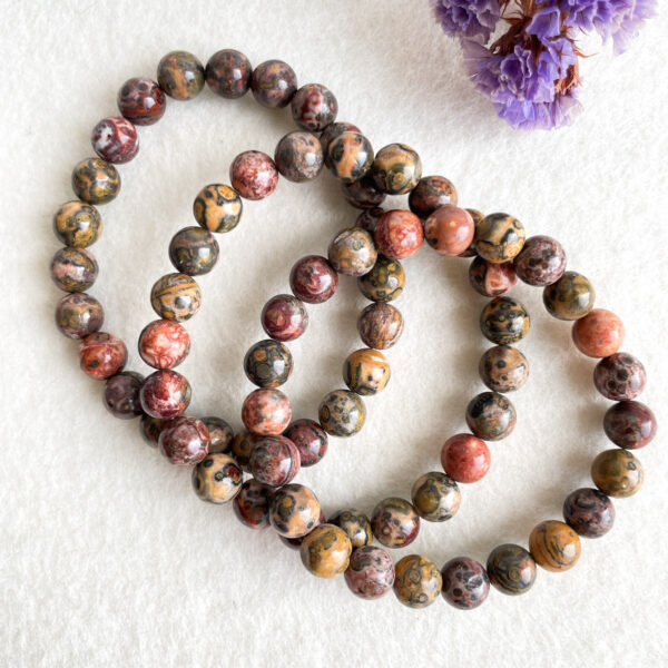 A string of colorful, patterned beads arranged in a spiral on a white textured background, with purple dried flowers in the upper right corner.