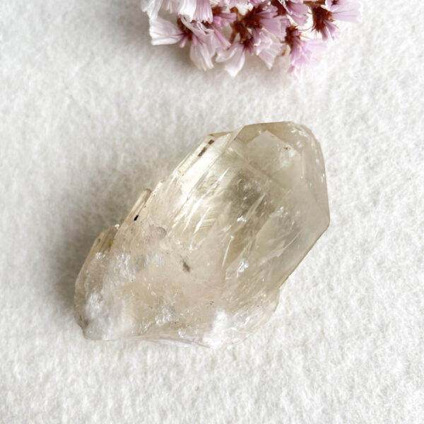A transparent crystal with naturally faceted surfaces on a white textured background, accompanied by a small cluster of pink flowers in the corner.