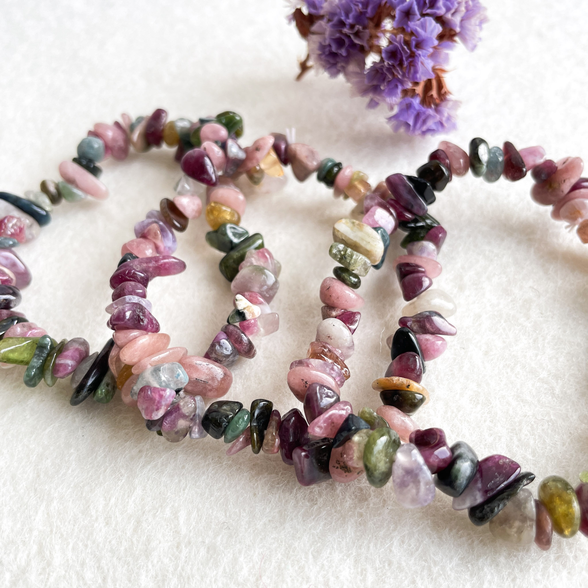 A beaded bracelet made of various colors of tourmaline strands on a white textured background, with a bundle of dried purple flowers in the upper right corner.