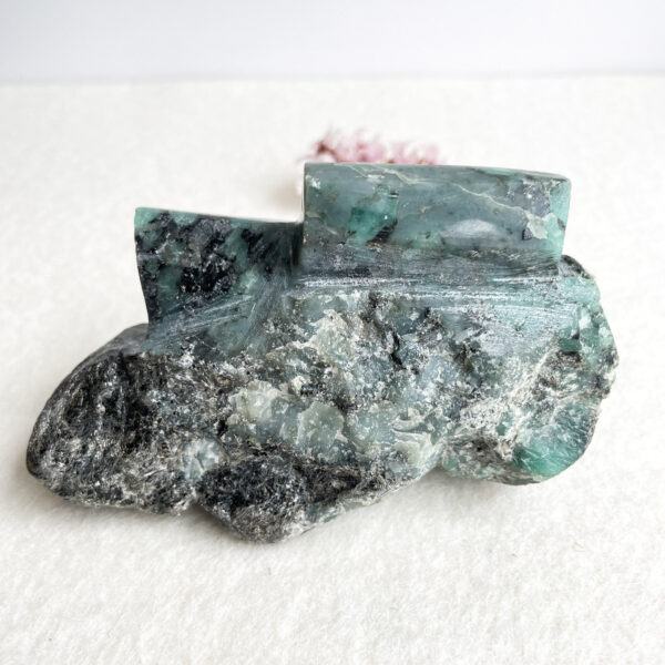 A polished piece of blue-green fluorite with rough edges and crystalline textures, on a pale surface with a blurred pink flower in the background.