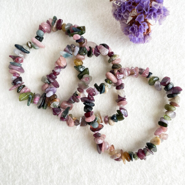 Two bracelets made of multicolored, tumbled gemstones are laid out in the shape of a heart. In the top right corner, there is a small bunch of dried purple flowers against a white textured background.