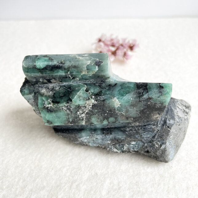 A polished green emerald crystal atop a natural uncut stone with a white background and a small pink flower in the background to the right.