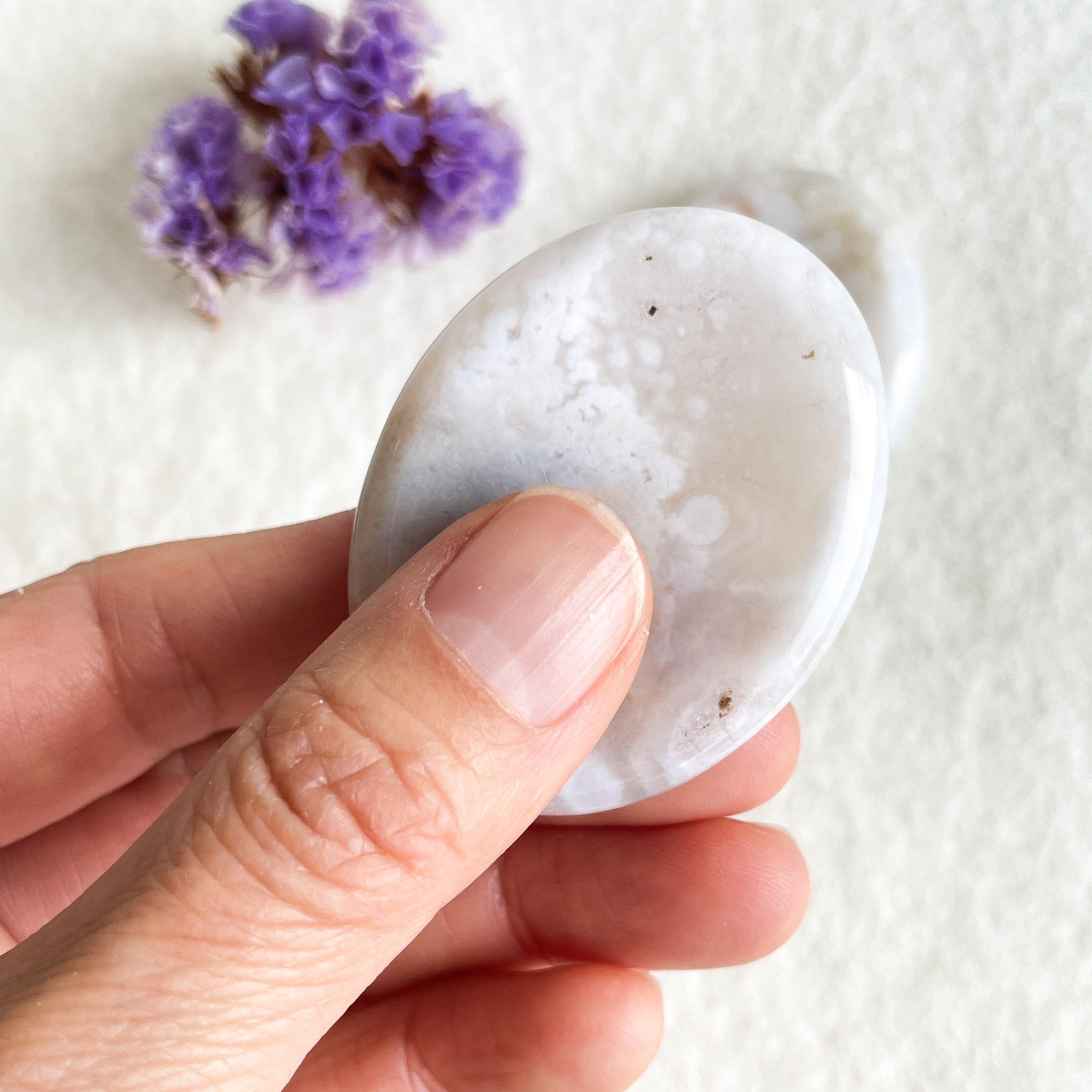 A close-up of a hand holding a smooth, polished white agate stone with natural patterns, next to small purple flowers on a textured white background.