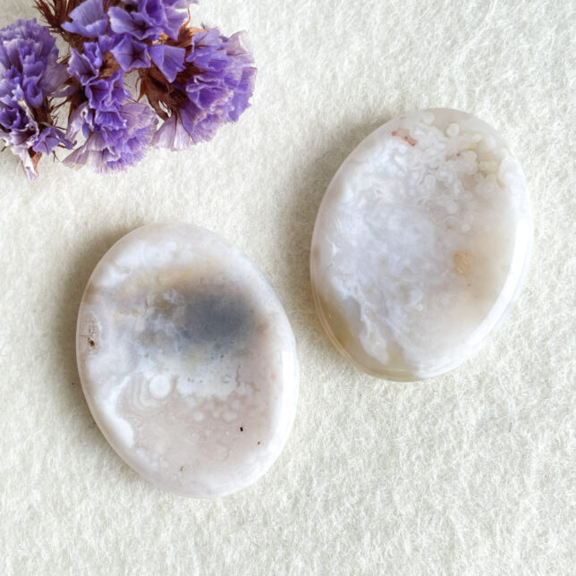 Two polished oval agate stones on a textured surface with dried purple flowers in the top left corner.