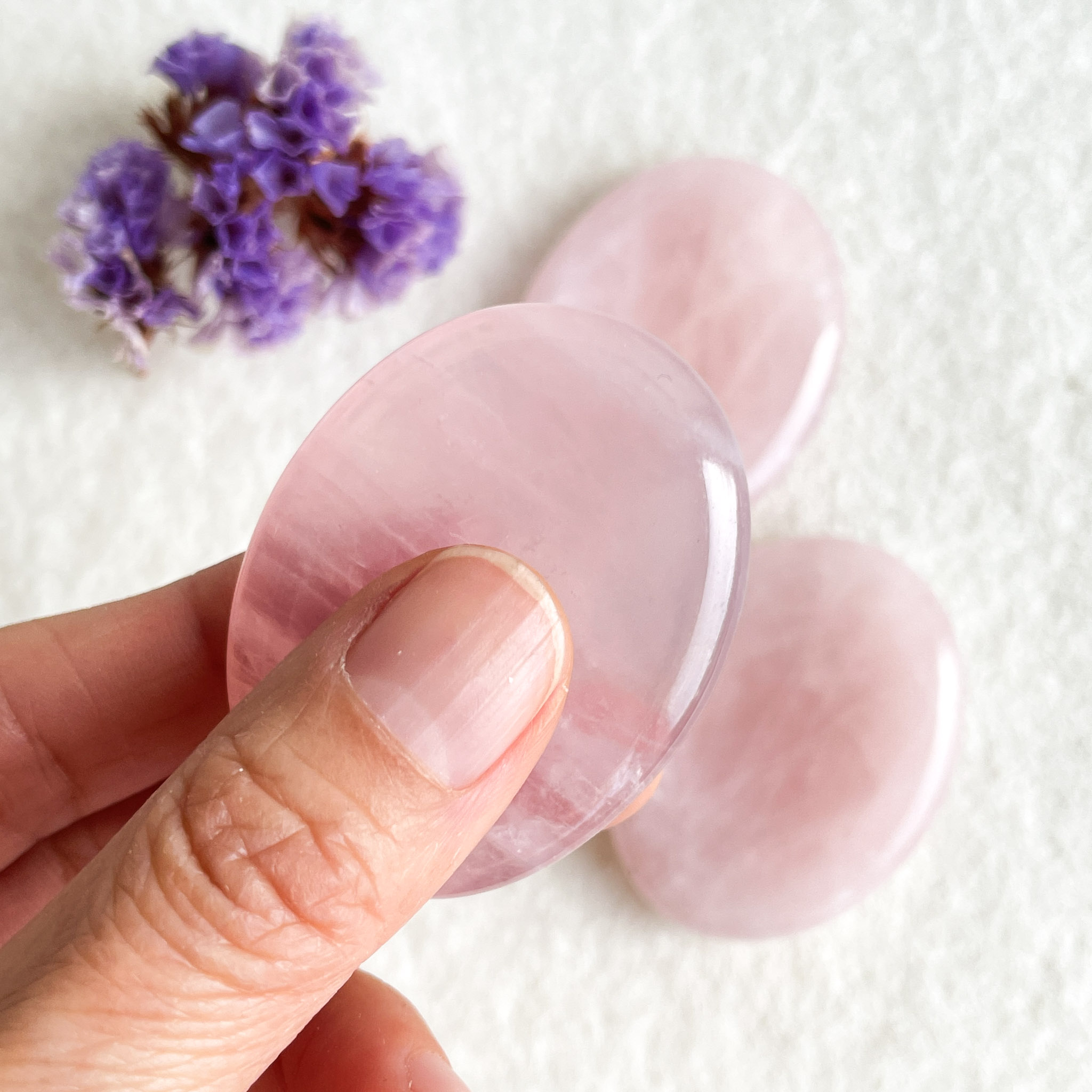 A hand holding a translucent pink crystal with two similar crystals and a bunch of small purple flowers in the background on a white surface.