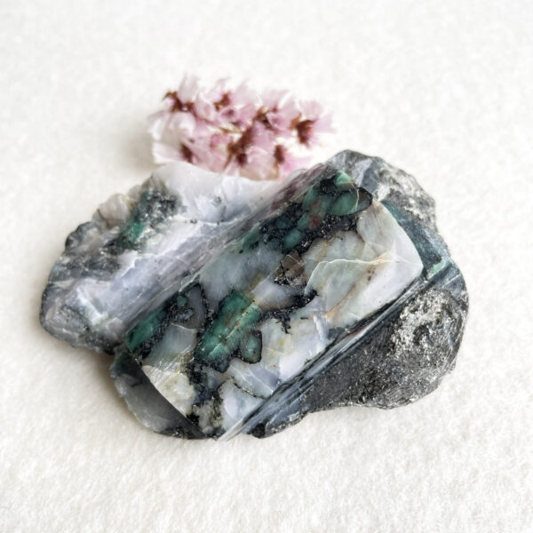 A polished cross-section of banded agate with varying shades of green, white, and gray, beside a small cluster of delicate pink flowers, on a textured white background.