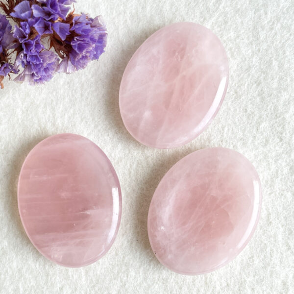 Three polished rose quartz stones on a textured white background with a cluster of dried purple flowers on the upper left side.